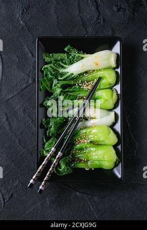 Cooked bok choy with sesame seeds and chili pepper olive oil in black square ceramic plate with black chopsticks over black textured background. Flat Stock Photo