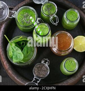 Opened glass jars and bottle of green spinach smoothie, served with baby spinach leaves, chia seeds, honey and lime in dark clay tray over old wood te