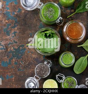 Opened glass jars and bottle of green spinach smoothie, served with baby spinach leaves, chia seeds, honey and lime over old wood textured background.