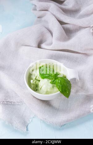 Homemade basil ice cream with fresh basil leaves in white ceramic bowl on white textile rag over blue textured background. Stock Photo