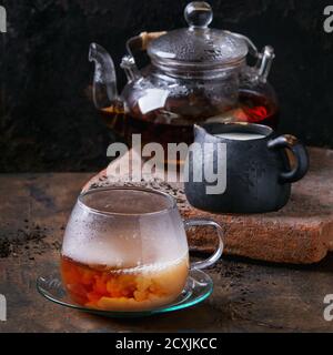 Glass cup on saucer of black tea with milk, teapot with hot black tea, standing on clay board with jug of milk over old wooden background. With copy s Stock Photo