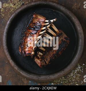 Whole grilled bbq rack of lamb, served with seasoning on clay tray over old wooden background. Overhead view. Square image Stock Photo