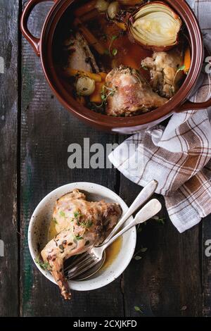 Brown ceramic pot with stewed rabbit with bouillon, vegetables and herbs, and bowl with rabbit leg, served with kitchen towel and vintage cutlery over Stock Photo