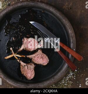 Chopped grilled bbq rack of lamb, served with seasoning, fork and knife on clay tray over old wooden background. Overhead view. Square image Stock Photo