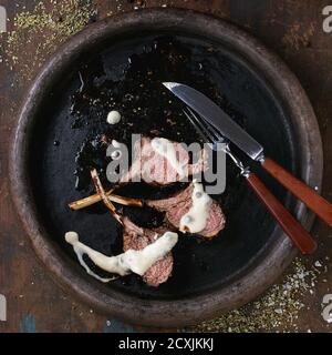 Chopped grilled bbq rack of lamb under pepper sauce,, served with seasoning, fork and knife on clay tray over old wooden background. Top view. Square Stock Photo