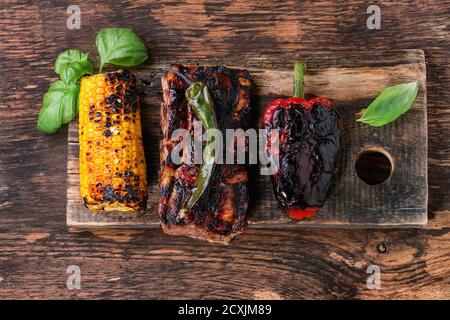 BBQ grilled pork ribs, corn, green chili and red bell pepper, served on wooden chopping board with fresh basil over wooden background. Flat lay Stock Photo