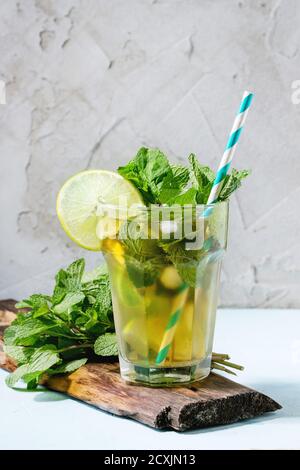 Glass of Iced green tea with lime, lemon, mint and ice cubes on wooden chopping board over light blue textured background. Stock Photo