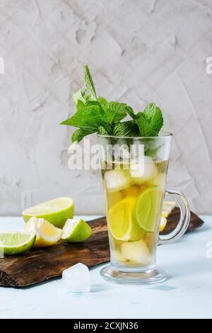 Glass of Iced green tea with lime, lemon, mint and ice cubes on wooden chopping board over light blue textured background. Stock Photo