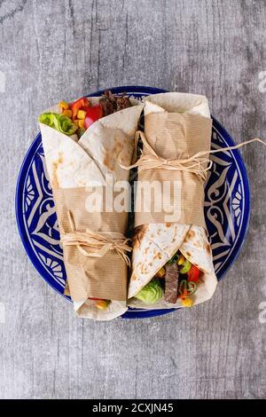 Mexican style dinner. Two papered tortillas burrito with beef and vegetables served on blue ornamental ceramic plate over white wooden background. Fla Stock Photo