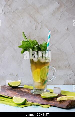 Glass of Iced green tea with lime, lemon, mint and ice cubes on wooden chopping board over light blue textured background with green textile napkin. Stock Photo