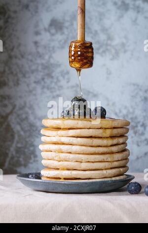 Pancakes with fresh blueberries in blue ceramic plate, and flowing honey from wooden honey dipper, served on white tablecloth with blue textured backg Stock Photo