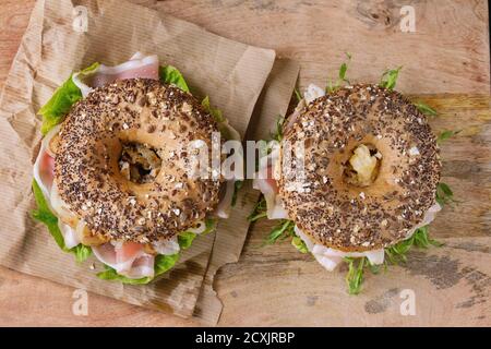 Two Whole Grain bagels with fried onion, scrambled eggs, green salad and prosciutto ham on paper over wooden textured background. Top view Stock Photo
