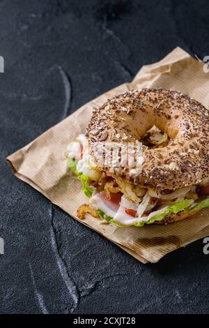 Sprinkle seeds Whole Grain bagel with fried onion, green salad and prosciutto ham on paper over black stone textured background. Stock Photo