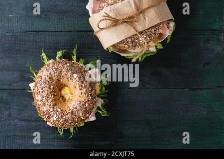 Two sprinkle seeds Whole Grain bagels with fried onion, scrambled eggs, green salad and prosciutto ham over dark wooden textured background. Top view. Stock Photo