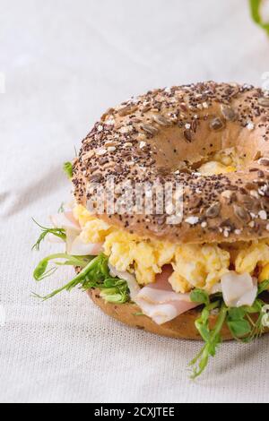 Whole Grain bagels with scrambled eggs, pea sprout and prosciutto ham on wood plate over wooden textured background. Stock Photo