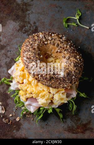 Opened Whole Grain bagels with scrambled eggs, pea sprout and prosciutto ham over old rusty iron textured background. Top view Stock Photo