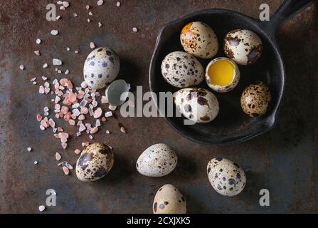 Whole and broken quail eggs with yolk in shell and pink sea salt crystal in small iron cast pan over old rusty texture metal background. Top view with Stock Photo