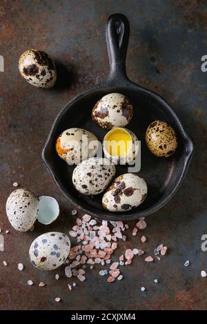 Whole and broken quail eggs with yolk in shell and pink sea salt crystal in small iron cast pan over old rusty texture metal background. Top view with Stock Photo