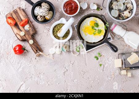 Breakfast with fried quail eggs in iron cast pan, cherry tomatoes, onion, ketchup sauce, seasonings in mortar, cheese grater, egg shell in pot. Beige Stock Photo