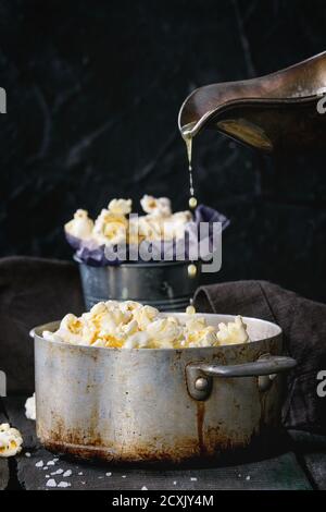 Prepared buttered popcorn served with sea salt in small buckets and vintage aluminum pan on old wooden kitchen table. Melted butter is poured from a s Stock Photo
