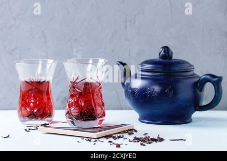 Two traditional asian style glasses with hibiscus tea karkade, served with dry hibiskus and dark blue ceramic teapot over blue and gray textured backg Stock Photo