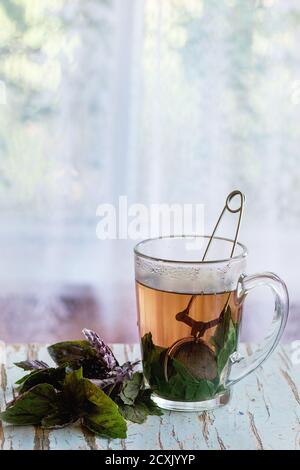 Glass cup of hot herbal tea with bunch of fresh violet basil, served with vintage tea-strainer on old wooden stool with window at background. Rustic s Stock Photo