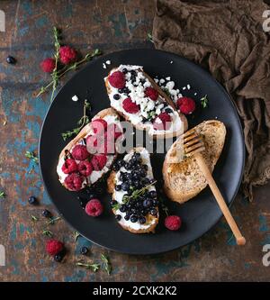 Set of dessert sandwiches with fresh berries blueberry and raspberry, cream cheese, thyme and honey, served with honey dipper on black plate over old Stock Photo