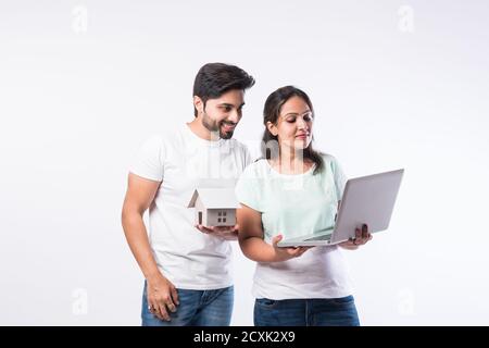 Indian young family couple choosing new home online, search real estate to buy or rent, house for sale on screen of computer, standing against white b Stock Photo