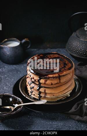 Stack of homemade american ombre chocolate pancakes with carob honey sauce served on black plate with jug of cream and teapot over black stone texture Stock Photo