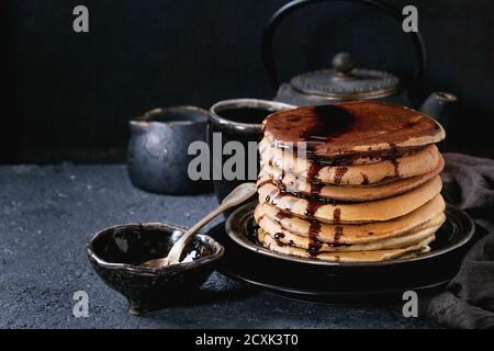 Stack of homemade american ombre chocolate pancakes with carob honey sauce served on black plate with jug of cream and teapot over black stone texture Stock Photo