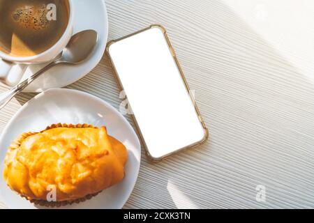 Flat lay, empty smartphone on table with hot coffee and snack or bakery with morning light. Top view image with empty space for copy and design. Clipp Stock Photo