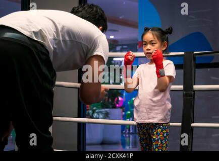 7 years old child girl learning Thai boxing in gym. Asian child in act of boxing guard, listen to coach teaching. Standing on boxing ring. Stock Photo