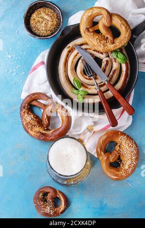 Fried sausage in iron-cast pan with fork and knife, glass of lager beer, mustard and pretzels on white kitchen towel over blue wooden background. Top Stock Photo