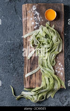 Fresh raw uncooked homemade green spinach pasta tagliatelle with egg yolk and flour on wooden chopping cutting board over dark texture concrete backgr Stock Photo