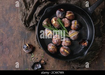 Roasted chestnuts in the ashes with rosemary in cast-iron pan on sackcloth over old dark wooden background. Top view with space for text. Stock Photo