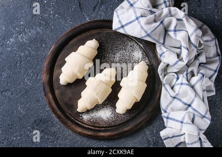 Raw unbaked croissant on wooden chopping board with kitchen towel over black texture concrete background. Top view Stock Photo
