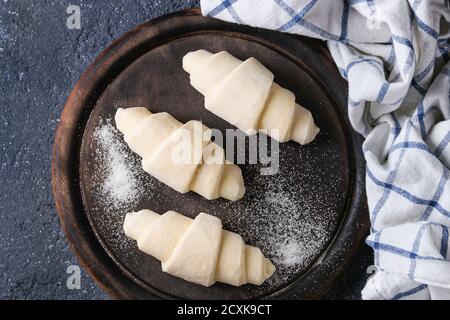 Raw unbaked croissant on wooden chopping board with kitchen towel over black texture concrete background. Top view Stock Photo