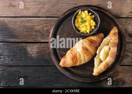 Breakfast with two croissant, butter and sliced mango fruit, served on wood round serving board over old wooden background. Top view with space. Stock Photo