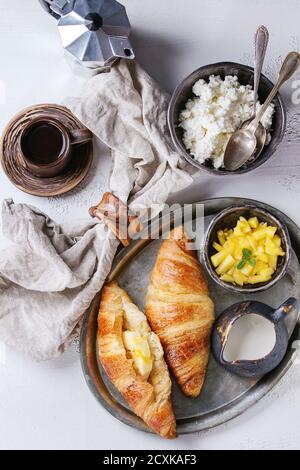 Breakfast with two croissant, butter, cup of coffee, cottage cheese and sliced mango fruit, served on serving metal tray with textile napkin on white Stock Photo