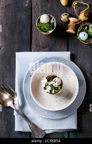 Table setting decor colorful Easter quail eggs with spring cherry flowers, moss in garden pots, empty plates, cutlery over old wood background. Dark r Stock Photo