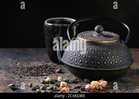 Black iron teapot with ceramic cup of hot tea and variety of black, green and herbal dry tea leaves and rose buds above over old dark wooden backgroun Stock Photo