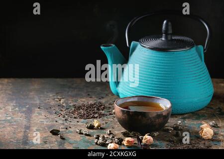 Turquoise iron teapot with ceramic cup of hot tea and variety of black, green and herbal dry tea leaves and rose buds above over old dark wooden backg Stock Photo