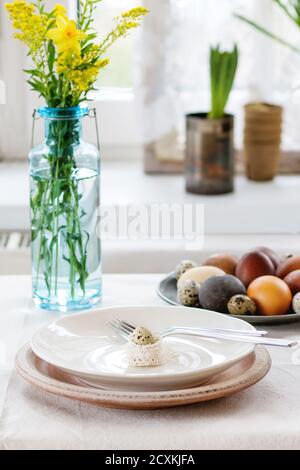 Prepare to Easter. White table setting with empty plates and fork on linen tablecloth decorated by colored brown chicken and quail eggs with yellow fl Stock Photo