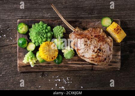 Grilled veal tomahawk steak with vegetables brussels sprouts, romanesco and corn cobs served on wooden serving chopping board over old wood background Stock Photo