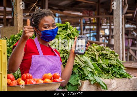 african woman selling in a local market wearing a face mask, holding a pos machine gives a thumbs up Stock Photo