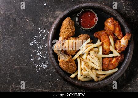 Fast food fried crispy and spicy chicken legs, wings and french fries potatoes with salt and ketchup sauce served in stone plate over dark texture bac Stock Photo