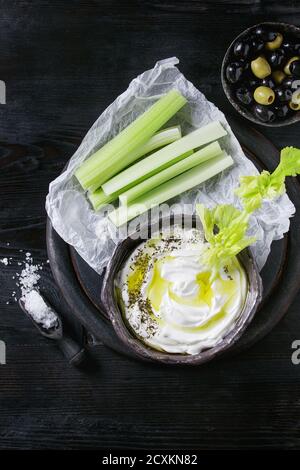 Fresh celery with yogurt and olive oil dip in ceramic bowl, served with sea salt and black, green olives on wood chopping board over black wooden burn Stock Photo