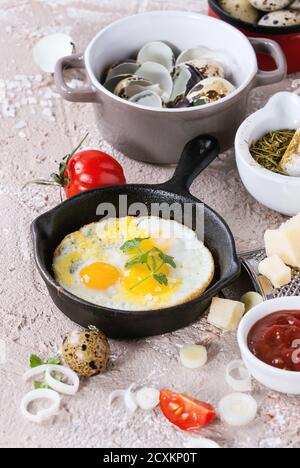 Breakfast with fried quail eggs in iron cast pan, cherry tomatoes, onion, ketchup sauce, seasonings in mortar, cheese grater, egg shell in pot. Beige Stock Photo