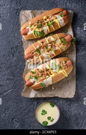 Hot dogs with sausage, fried onion, coriander leaves, cheese sauce and mustard, served on baking paper over black concrete texture background. Fast fo Stock Photo