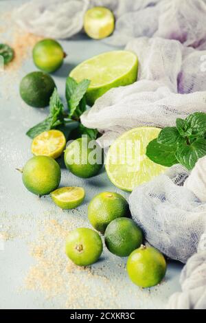 Ingredients for mojito cocktail, whole, sliced lime and mini limes, mint leaves, brown crystal sugar over gray stone texture background with gauze tex Stock Photo
